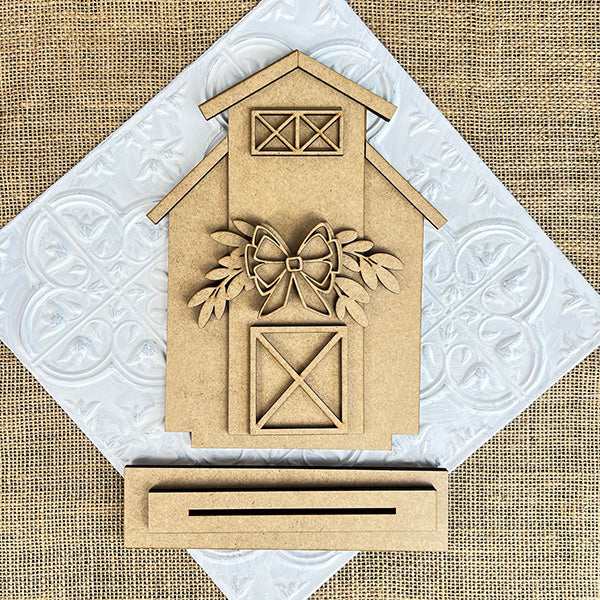 The Barn Shelf Sitter from Janet's Craft Corner exudes rustic charm with its bow and leaves design on a decorative white tile. The burlap fabric background enhances the cozy elegance of this wooden barn-shaped ornament, part of a DIY seasonal home decor craft kit.