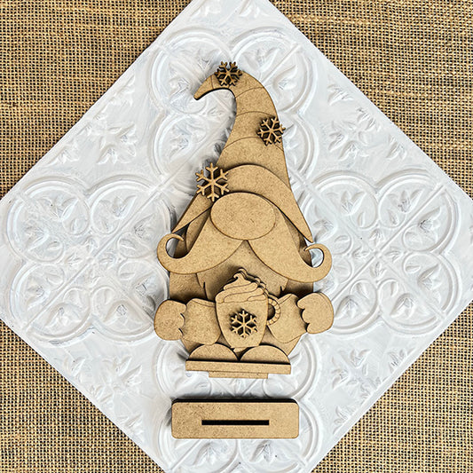 The Hot Cocoa Gnome from Janet's Craft Corner features a tall hat, bushy beard, and snowflake mug. This DIY shelf sitter kit offers seasonal home decor with a textured white diamond on a woven beige background.