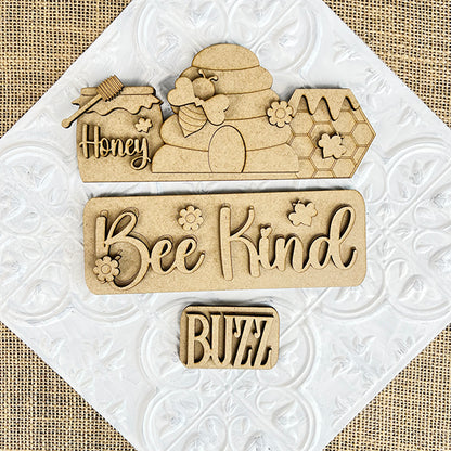 The *Bee Interchangeable Insert* by Janet's Craft Corner is a hand-painted DIY home decor kit featuring wooden cutouts on a textured background. It includes designs of bees, honeycombs, and a beehive, alongside charming phrases such as "Honey," "Bee Kind," and "Buzz." This kit is perfect for refreshing your space with interchangeable decor.