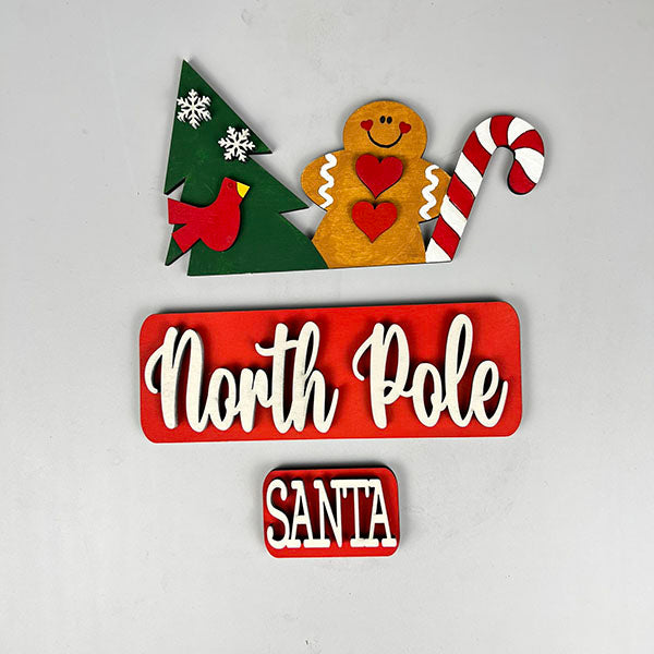 Set against a white background, the festive decorations feature a hand-painted DIY craft kit from Janet's Craft Corner. It includes a Christmas tree with snowflakes, a red bird, and an interchangeable North Pole insert. Completing this charming home decor set are a gingerbread figure and candy cane.