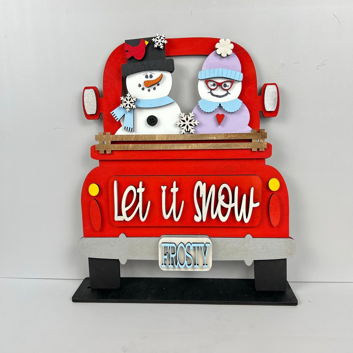 The "Let It Snow" Interchangeable Insert DIY home decor kit from Janet's Craft Corner showcases a charming red truck decoration featuring a snowman couple and a winter-clad woman in the back. The truck, adorned with "Let it Snow" lettering and a "Frosty" license plate, highlights figures wearing cozy winter hats and scarves, making it perfect for your own DIY craft project masterpiece.