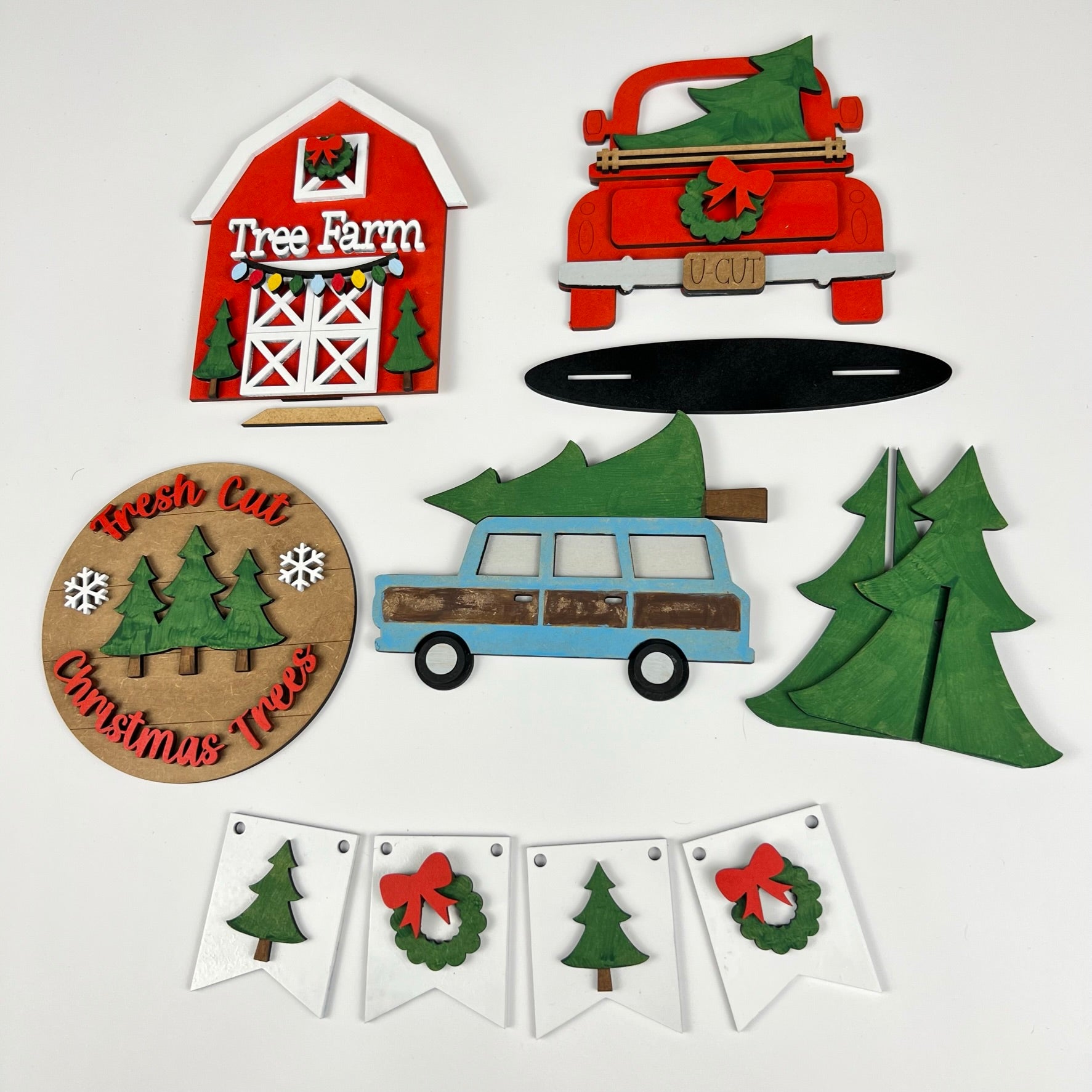 The Tree Farm - DIY Tiered Tray Kit from Janet's Craft Corner includes Christmas-themed wooden decorations such as a red barn with "Tree Farm" signage, a red and blue car, trees, a circular "Fresh Cut Christmas Trees" sign, and a festive banner decorated with trees and wreaths.