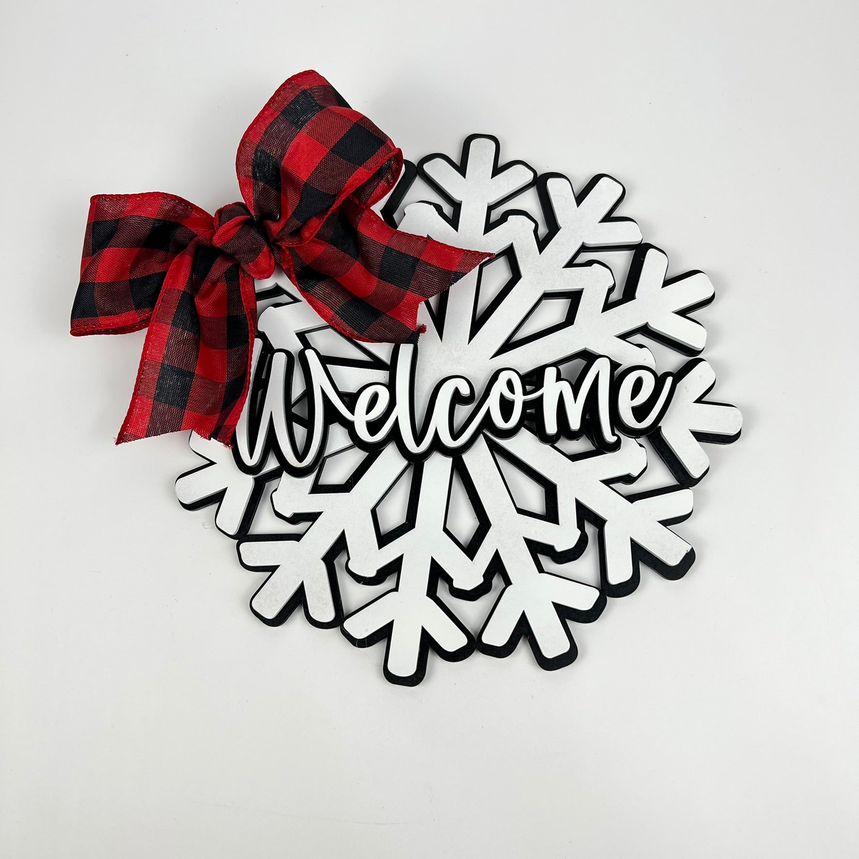 The Welcome Snowflake Door Hanger by Janet's Craft Corner showcases a snowflake design with "Welcome" in the center. A red and black checkered bow at the top left adds a festive touch, while rustic wood accents bring warmth against the plain white background.