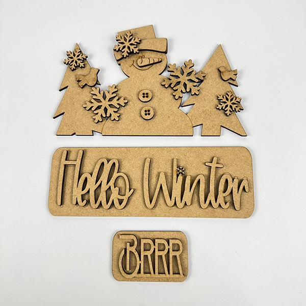 Add a festive touch to your space with Janet's Craft Corner's DIY home decor kit, the "Hello Winter Interchangeable Insert." This kit features wooden cutouts of a snowman in a hat, trees, and snowflakes. It includes pieces that read "Hello Winter" and "Brrr," making it perfect for changing up your home decor on any plain background.