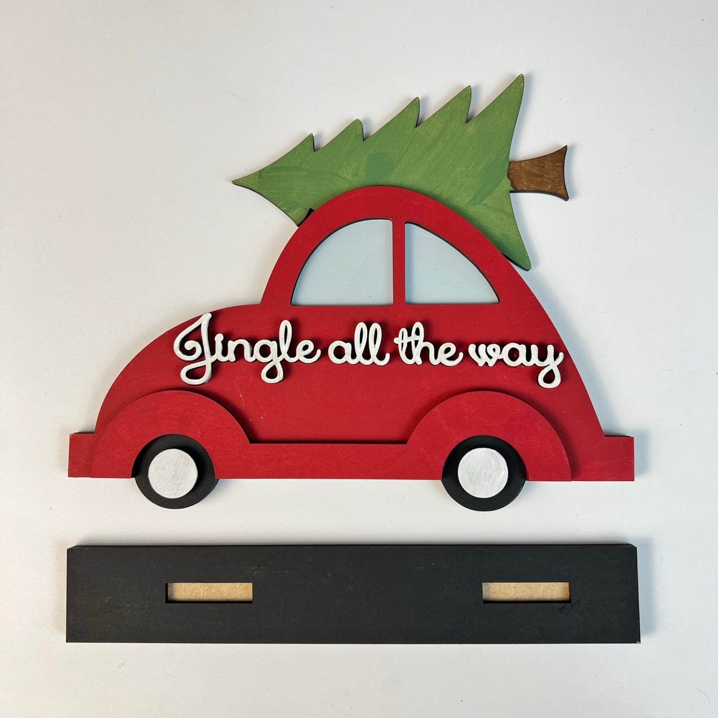 Janet's Craft Corner offers the Jingle All the Way Volkswagen Shelf Sitter, a wooden decoration featuring a red Volkswagen car adorned with a green evergreen tree on top. This festive piece, complete with the phrase "Jingle all the way" in white lettering, sits on a black rectangular stand equipped with two slots for seamless display.