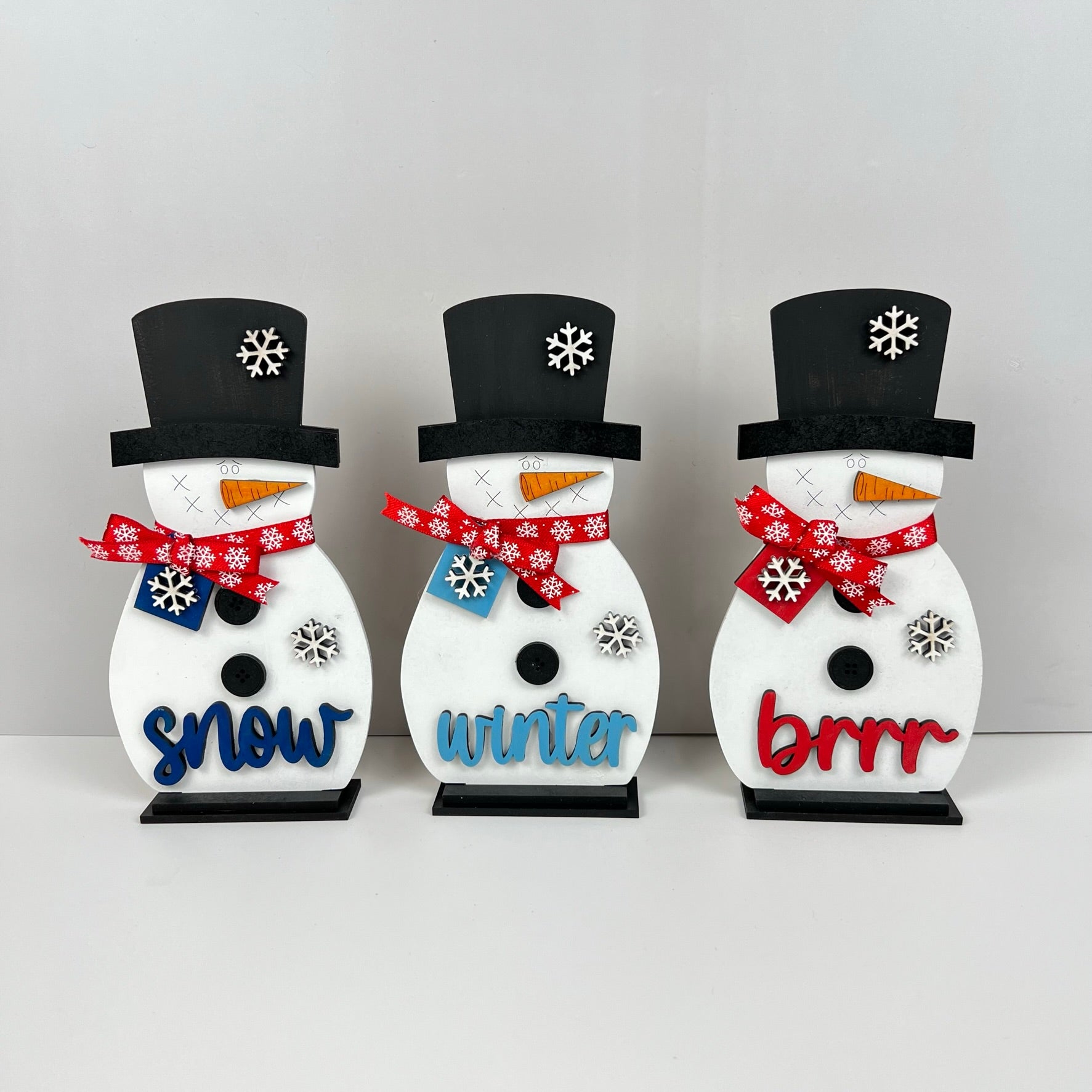 A delightful trio of Snowman Shelf Sitters from Janet's Craft Corner, each adorned with black hats and red scarves, featuring a charming snowflake design. The festive words "snow," "winter," and "brrr" are beautifully emblazoned in blue and red across their bodies. Perfect for adding charm to your home décor, they stand gracefully against a plain white background.