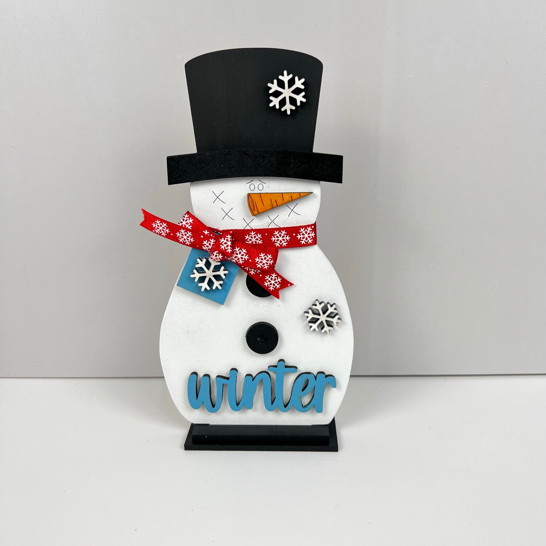 This delightful Snowman Shelf Sitter from Janet's Craft Corner is ideal for enhancing your home décor. The figurine showcases a black top hat, carrot nose, red scarf, and black buttons embellished with snowflakes. Adding to its charm, the word "winter" is gracefully inscribed in blue across the base, bringing a festive ambiance to any room.