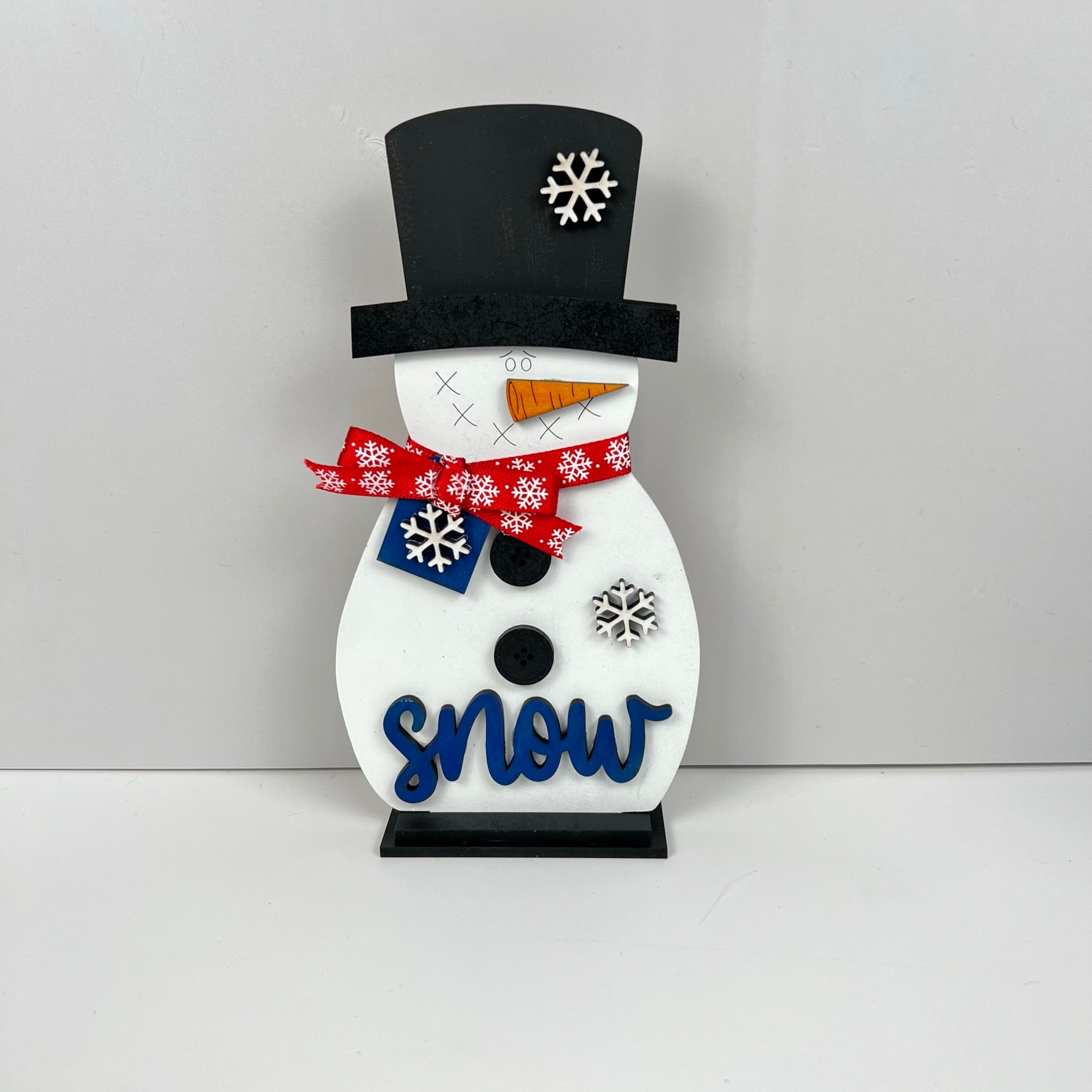 Enhance your home décor with Janet's Craft Corner Snowman Shelf Sitter. Sporting a black top hat, a red scarf embellished with white snowflakes, and two black buttons, this charming snowman features a carrot nose and the word "snow" in blue against a simple gray backdrop.