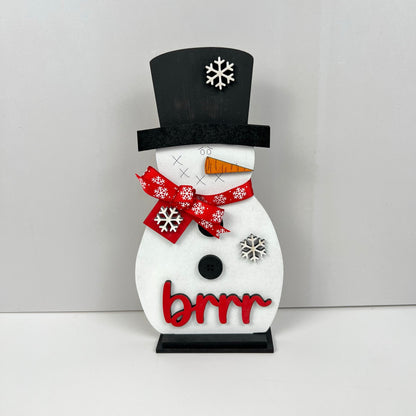 Introducing the Snowman Shelf Sitter by Janet's Craft Corner! This charming decorative piece features a black top hat adorned with white snowflakes, a red bow highlighted with snowflakes, and a carrot nose. Ideal for home decor, it is further enhanced by the word "brrr" in bold red letters.