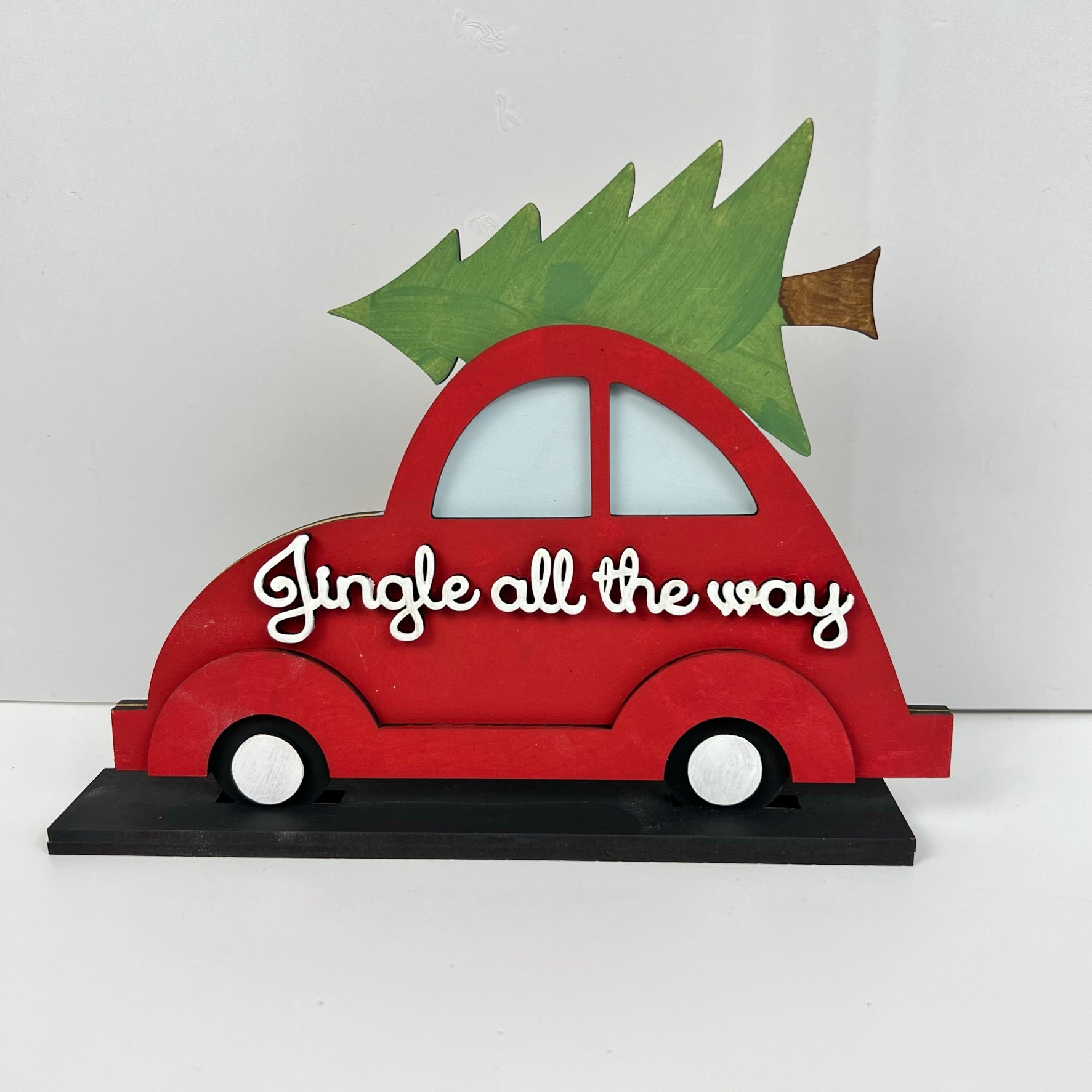 The Jingle All the Way Volkswagen Shelf Sitter by Janet's Craft Corner is a decorative piece featuring a red Volkswagen car with an evergreen tree on top, embellished with the phrase "Jingle all the way" in white script, perfectly capturing the spirit of yuletide cheer. The car is showcased against a plain white backdrop.