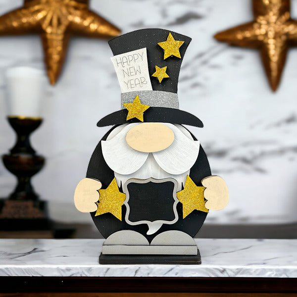 The Happy New Year Gnome Shelf Sitter from Janet's Craft Corner sits on a marble surface, wearing a top hat with gold stars. Blurred star-shaped ornaments enhance the cozy and inviting New Year's decor scene.