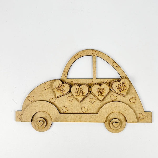 Janet's Craft Corner offers a Valentine Volkswagen Shelf Sitter DIY kit, featuring a vintage-style car adorned with heart shapes, some engraved with messages like "BE MINE" and "KISS ME," adding charm to this seasonal home decor.