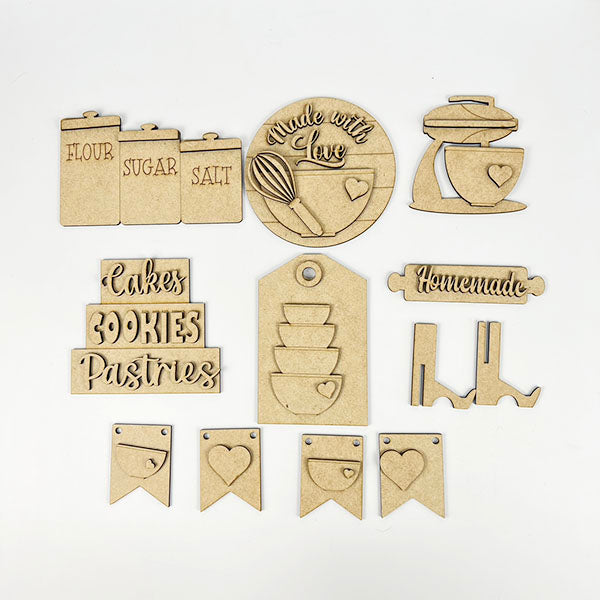 The Baking Decor Set by Janet's Craft Corner offers a charming farmhouse collection of 7 kitchen-themed wooden cutouts: labeled jars ("flour," "sugar," "salt"), a whisk circle ("made with love"), a baking mixer, words like "cakes" and "cookies," bowls, and flags with hearts.