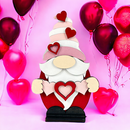 Introducing the Valentine Gnome Shelf Sitter from Janet's Craft Corner, a charming addition to your home décor. This wooden gnome figurine is dressed in red and pink, adorned with heart decorations. Set against a pink background, it is encircled by vibrant red and pink heart-shaped balloons.