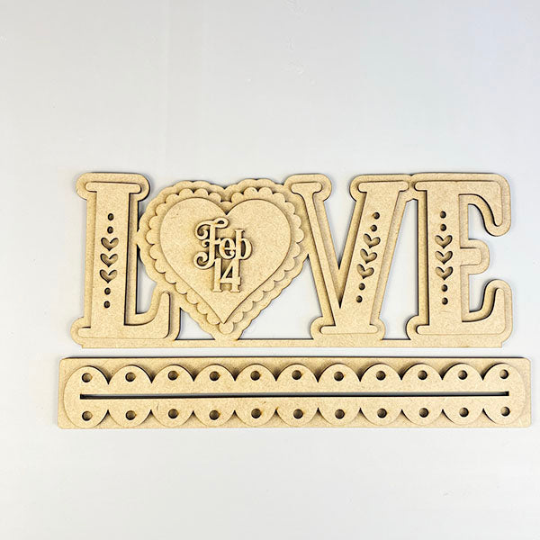 The "Love Shelf Sitter" DIY kit from Janet's Craft Corner spells "LOVE" with a heart as the "O" and includes a "Feb 14" detail. Below is a separate piece adorned with small heart cutouts, ideal for personalizing your home décor.