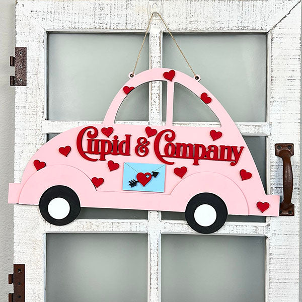 A decorative DIY door hanger from Janet's Craft Corner, in the shape of a pink Cupid Volkswagen, adorns a white door. The sign reads "Cupid & Company" in red letters, embellished with red hearts and an arrow through a heart emblem on the side—perfect for adding charm to your home décor kit.