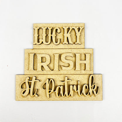 The "St Patricks Decor Set" by Janet's Craft Corner is a DIY kit featuring tiered tray wooden signs with the words "LUCKY," "IRISH," and "St. Patrick" in varied fonts on a light background, perfect for festive home decor.