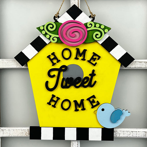 The Home Tweet Home Birdhouse Door Hanger by Janet's Craft Corner is a charming decorative piece featuring a yellow birdhouse sign with black and white striped roof edges. It is adorned with a pink rose and green leaves on top, making it the perfect addition to your home décor. The sign displays "Home Tweet Home" in black letters and includes a small blue bird in the bottom right corner.