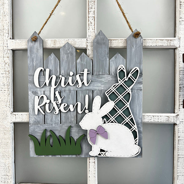 An Easter door hanger from Janet's Craft Corner, featuring "He is Risen" with two bunny cutouts—one plaid, one white with a purple bow—set against a gray picket fence background with green grass accents.