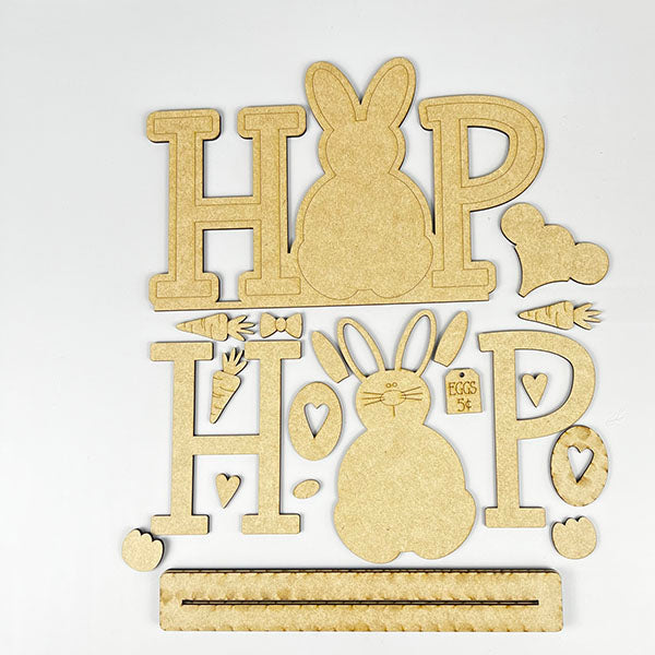 The HOP Standing Bunny Shelf Sitter by Janet's Craft Corner is a DIY decor kit featuring "HOP" with a bunny silhouette as "O." It includes cutouts like carrots, hearts, and eggs, and a slotted rectangular piece for easy assembly—ideal for holiday projects.