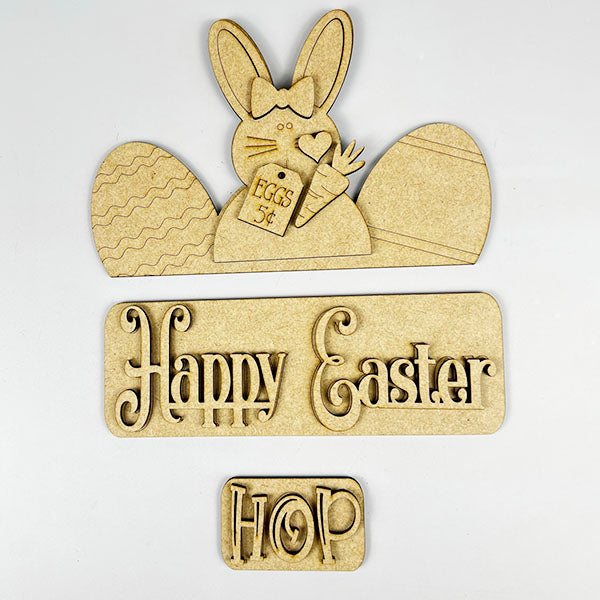 This Easter Bunny Interchangeable Insert from Janet's Craft Corner is a delightful wooden decoration kit, featuring an Easter Bunny holding a "Eggs 5¢" sign and surrounded by two egg shapes. The words "Happy Easter" and "Hop" are intricately carved beneath in decorative letters, making it ideal for DIY craft enthusiasts or as versatile home decor.