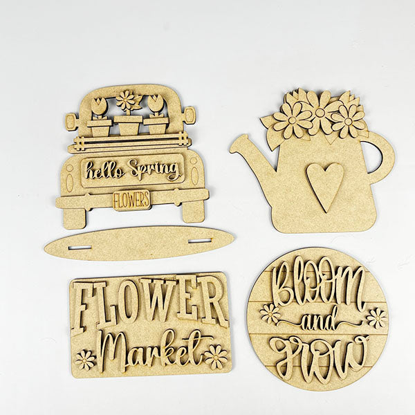 Janet's Craft Corner offers a Flower Market Mini Shelf Sitters set, featuring wooden cutouts of a truck with potted plants labeled "hello Spring," a watering can with flowers, and signs that read "Flower Market" and "Bloom and Grow," perfect for your DIY spring tiered tray decor.