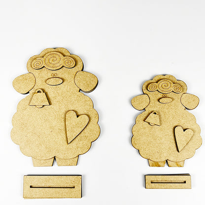 Janet's Craft Corner offers a Lamb Shelf Sitter DIY Kit featuring two charming wooden sheep cutouts with curly wool, facial details, a heart on each body, and bottom slots. One is larger than the other against a white background—ideal as home décor!.