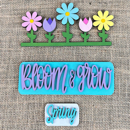 The Spring Blooms Interchangeable Insert from Janet's Craft Corner features colorful wooden cutouts on burlap. The top showcases pastel flowers, the middle highlights "Bloom & Grow" in purple on a teal background, and the bottom displays "Spring" in white against teal. It's perfect for home décor or as a hand-painted DIY craft kit.