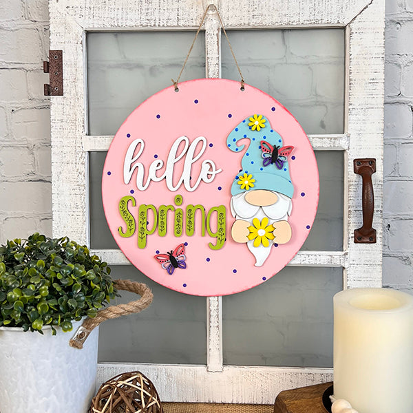 Set within a window frame, the Hello Spring Gnome Door Hanger from Janet's Craft Corner displays the cheerful "Hello Spring" text alongside a gnome in a blue hat embellished with flowers and butterflies. Complementary décor includes a plant, candle, and wicker ball set against a white brick wall, rounding out this delightful DIY setup.