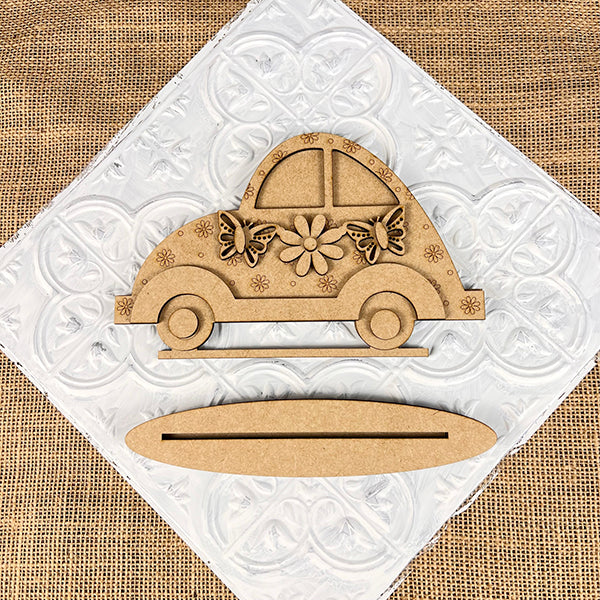 Janet's Craft Corner offers the Spring Volkswagen Shelf Sitter— a DIY decorative piece. This wooden car-shaped decor, featuring floral and butterfly designs, rests on a textured white square atop burlap with an oval wooden stand below, perfect for any home décor collection.