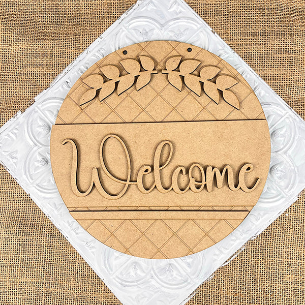 The Welcome Round Door Hanger by Janet's Craft Corner is a charming DIY home decor craft kit. This wooden sign features "Welcome" with leaf accents, a checkered pattern, and white textured surface over burlap, perfect for farmhouse decor.