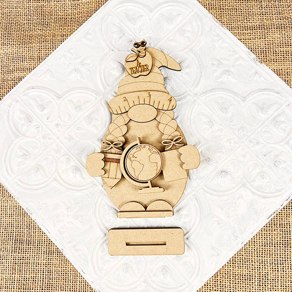 Introducing the Girl Teacher Gnome DIY Home Decor Craft Kit by Janet's Craft Corner. This charming wooden cutout features a gnome teacher holding a globe and books, with "Teacher" labeled on her hat. She stands on a textured white and burlap background, perfectly suited for home décor. Use the slot at the bottom to insert notes or photos, adding a delightful DIY craft element to any space.