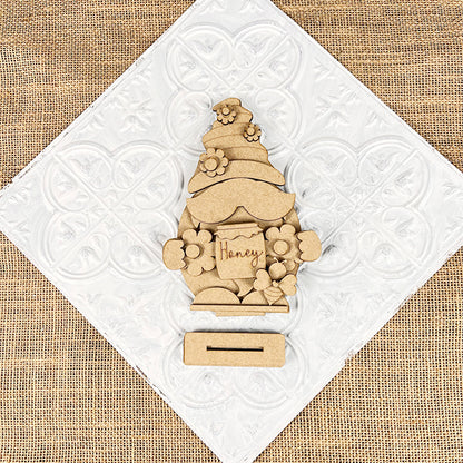 The Bee Gnome Shelf Sitter by Janet's Craft Corner is a charming DIY home decor kit. It features a flower-adorned figure holding a "Honey" sign, displayed on a white tile over burlap with an underside slot, perfect for farmhouse decor enthusiasts.