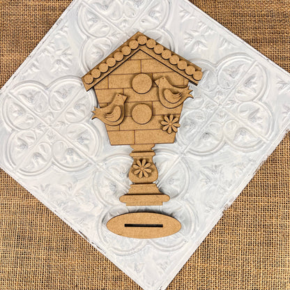 Janet's Craft Corner presents the Spring Standing Signs, a decorative DIY kit featuring a wooden birdhouse design against a white tile backdrop. The kit includes circular elements and two bird shapes, complemented by a floral motif at the base on textured burlap, making it ideal for home décor or as part of your Spring Standing Signs collection.