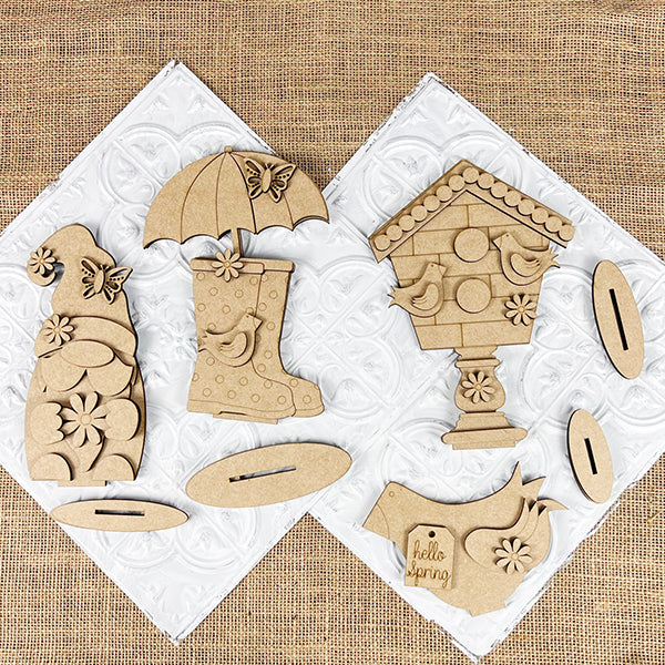 The Spring Mini Shelf Sitters by Janet's Craft Corner is a DIY decor kit including: a gnome with flowers, a rain boot with a butterfly, and birdhouses teeming with birds. "Hello spring" is etched on one bird. These pieces rest on textured surfaces above white tiles, perfect for your gnome décor collection.