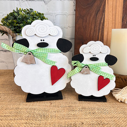 Incorporating two charming Lamb Shelf Sitters from Janet's Craft Corner, these white figurines with black ears and noses are displayed on a burlap surface. Each lamb is adorned with a green gingham bow featuring a bell and displays a red heart. Perfect for home décor, they create a cozy ambiance while nestled among plants against a backdrop of brick walls and wooden items.