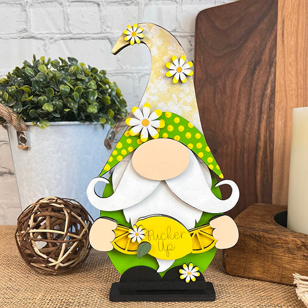 The Lemonade Gnome by Janet's Craft Corner, featuring a vibrant yellow polka dot hat and charming daisy accents, holds a lemon inscribed with "Pucker Up" and adds a delightful touch to your farmhouse decor. It stands beautifully on a table adorned with a small plant, woven ball, and candles in the background.