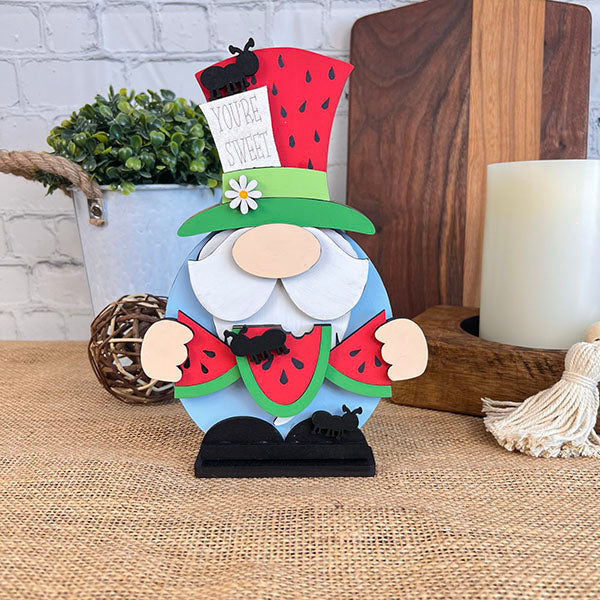 This delightful Watermelon Gnome from Janet's Craft Corner is ideal for farmhouse decor, showcasing a charming green and red hat with a "You're Sweet" sign. It holds watermelon slices and is embellished with tiny black ants. The scene includes a candle, plants, and rustic wooden pieces to create a cozy atmosphere.