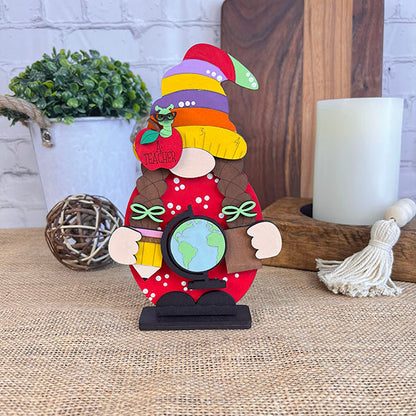 A delightful Girl Teacher Gnome from Janet's Craft Corner, adorned with a vibrant hat and a red polka dot outfit, holds a globe as she stands on a table. Accompanying her in this enchanting scene are a potted plant, wooden cutting board, and large white candle. A tassel and decorative ball add the perfect finishing touch to this charming home décor ensemble.