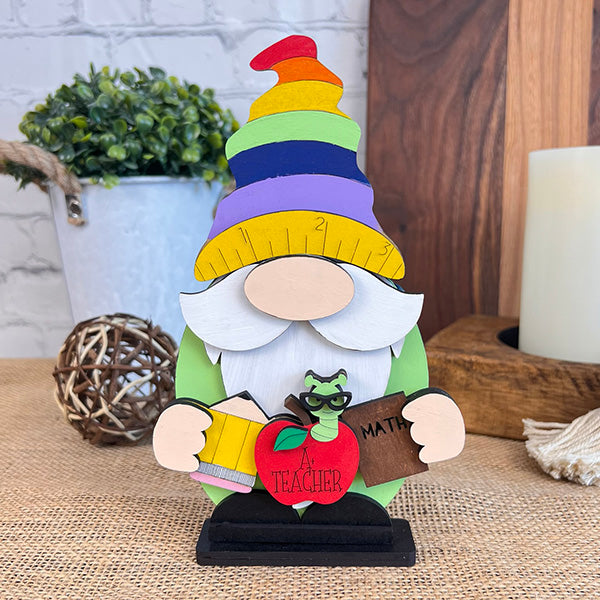 The Teacher Gnome Shelf Sitter by Janet's Craft Corner features a vibrant teacher gnome figurine with a tall, rainbow-colored hat, holding a book labeled "Math" and an apple inscribed with "Teacher." A cute green worm with glasses decorates the apple. Ideal for farmhouse decor, this charming scene is complemented by decorative items and a plant in the background.