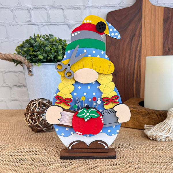 The whimsical Sewing - Quilting Gnome from Janet's Craft Corner, dressed in blue with white polka dots, showcases charming blonde braids and a sewing-themed hat embellished with buttons and scissors. This farmhouse-chic figure also holds a red pincushion, set against a backdrop of candles, plants, and rustic decor.