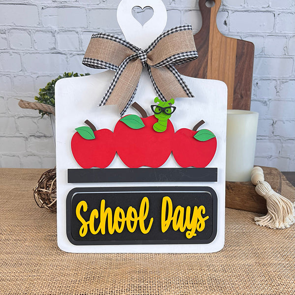 The School Days Interchangeable Insert by Janet's Craft Corner is a charming DIY home decor kit featuring a decorative sign adorned with three red apples and an adorable green worm wearing glasses. A black plaque below displays the words "School Days" in yellow, and it is completed with a black and white checkered bow accented with burlap, adding a touch of farmhouse decor charm.