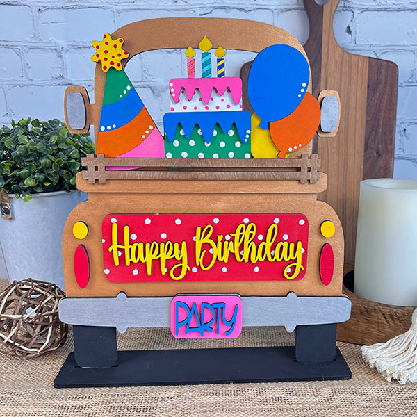 Introducing Janet's Craft Corner's DIY Home Decor Kit: The Antique Truck Base with Interchangeable Insert. This lively wooden truck display proudly showcases "Happy Birthday" on the back, complete with a party hat, balloons, and a layered cake design, perfectly suited for your festive occasions. It features an antique-style truck base that allows for customization with interchangeable inserts and is beautifully set against a rustic background alongside a small potted plant and charming decorative items.