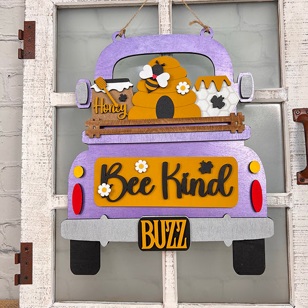 An antique truck base with an interchangeable insert from Janet's Craft Corner features a decorative purple wooden truck sign adorned with a beehive, honey jar, and flowers. Prominently displaying the words "Bee Kind" and "Buzz," this elegant piece is perfect for a hand-painted DIY home decor kit and looks great when hung on a white wooden door.