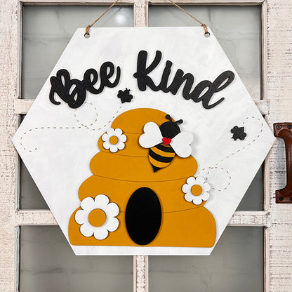 The Bee Kind Door Hanger from Janet's Craft Corner displays a hexagonal design showcasing the words "Bee Kind" at the top. It features a stylized hive and bee encircled by white flowers, making it an ideal addition to rustic farmhouse decor and bringing warmth to any wooden door with a vintage handle.