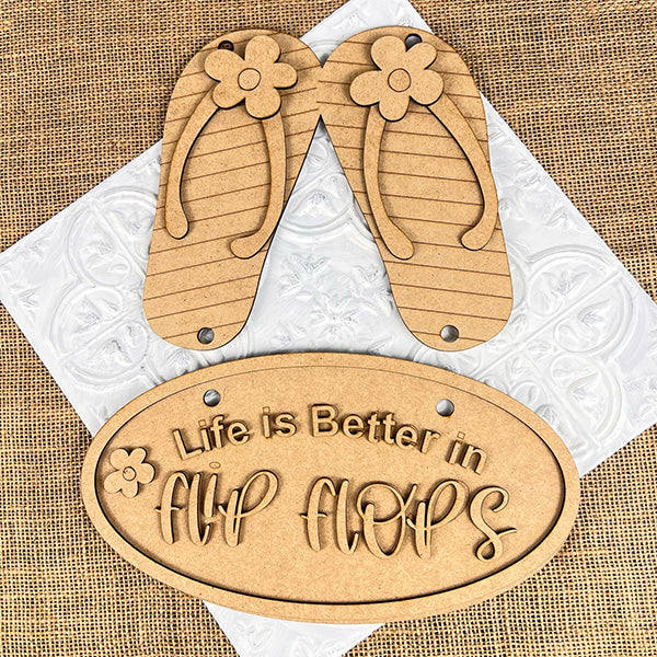 The Flip Flops Door Hanger from Janet's Craft Corner, set against a textured burlap background, features wooden cutouts and a floral decoration. An oval sign reads "Life is Better in Flip Flops," ideal for home décor or as part of the DIY seasonal craft kit.