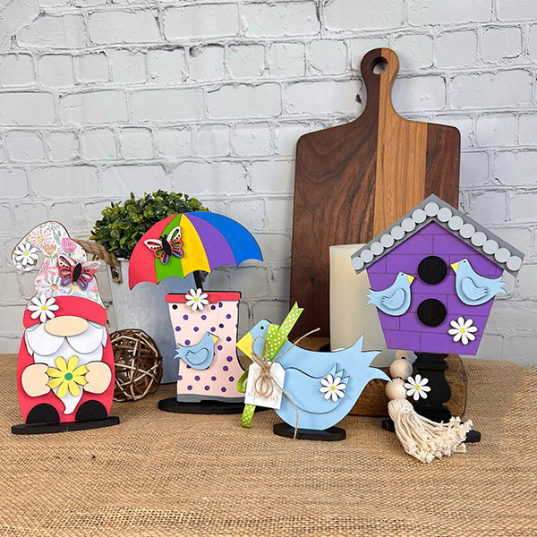 A delightful array of Janet's Craft Corner's Spring Standing Signs enhances the table, showcasing a gnome with a butterfly and a bird holding a polka dot umbrella. Adding to the charm, a birdhouse embellished with bluebirds completes the display. Perfect for home décor lovers, these pieces seamlessly integrate into any space.