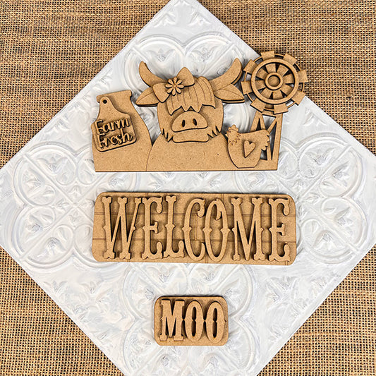 Janet's Craft Corner offers a delightful DIY home decor kit called the Farm Welcome Interchangeable Insert, perfect for adding farmhouse charm. This hand-painted set includes wooden cutouts featuring a cow with a windmill and a "Farm Fresh" tag, a "WELCOME" sign, and a small "MOO" sign. The burlap fabric background enhances the rustic appeal of this craft project.