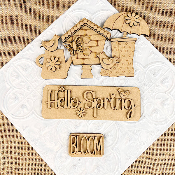 Janet's Craft Corner presents the "Hello Spring Interchangeable Insert" DIY home decor kit, featuring wooden cutouts of a birdhouse, birds, an umbrella, boots, and flowers on a textured background. The text reads "Hello Spring" and "BLOOM," making it perfect for crafting your own springtime decorative insert!