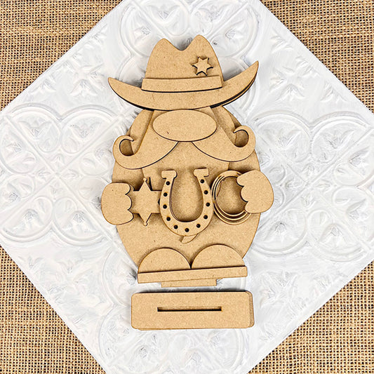 The Cowboy Gnome Shelf Sitter from Janet's Craft Corner is a DIY home decor craft kit that features a cowboy-themed gnome with hat, mustache, and boots. It holds a horseshoe and lasso against an embossed diamond panel on burlap, perfect for adding charm to your space.